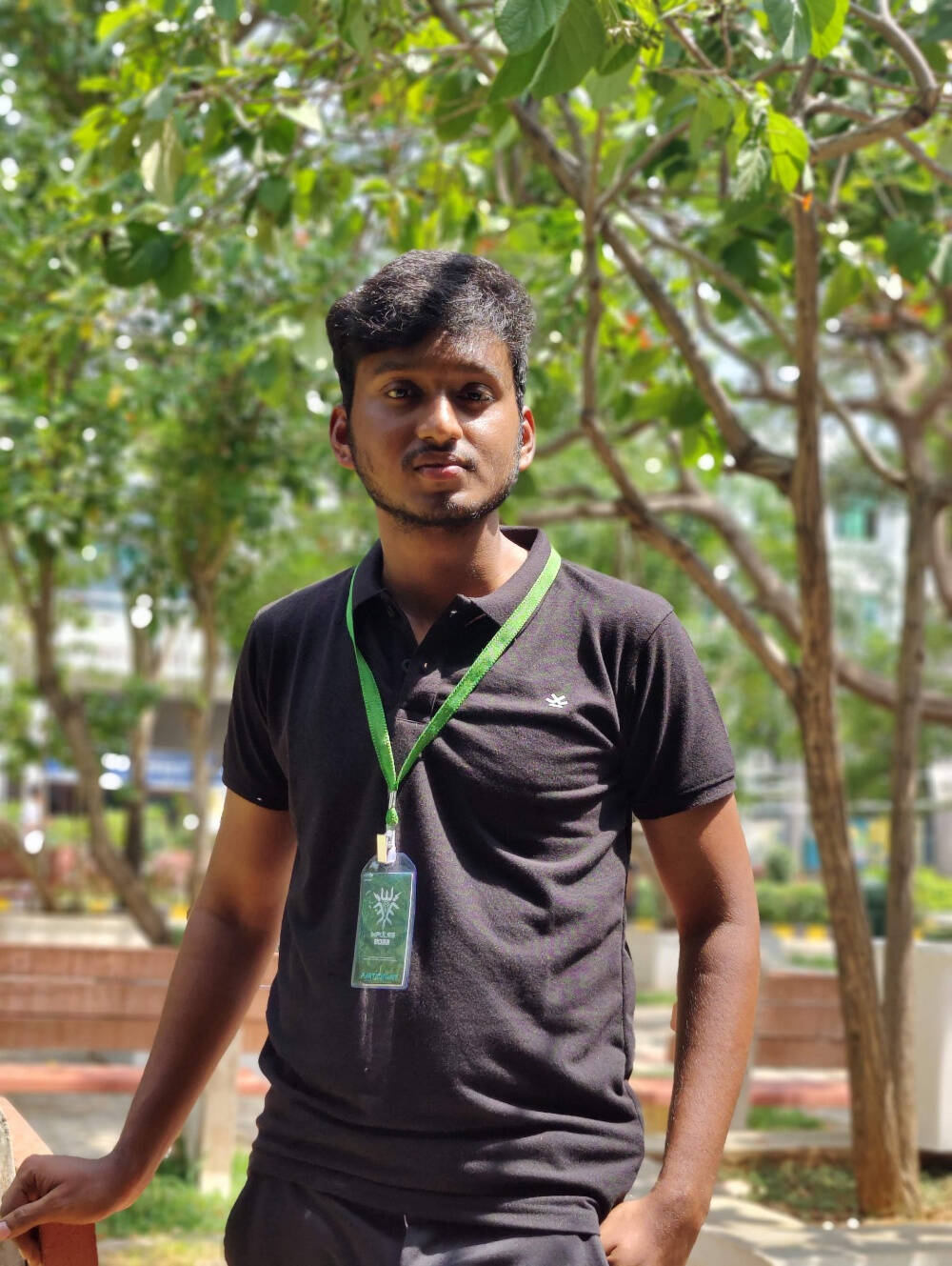 Sujish Kumar, wearing a dark polo shirt and a green lanyard with a badge, standing outdoors with a hand in pocket, surrounded by lush green trees and a park bench.